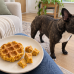 Generated illustration of a plain pumpkin waffle and cut pieces on a plate, with brindle Nina standing nearby in her living room.