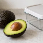 A halved avocado with its pit and a whole avocado on a kitchen countertop.