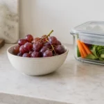 A bowl of grapes kept well back on a kitchen counter beside a lidded storage container.