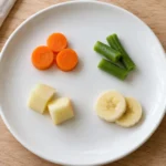 Small samples of carrot, green beans, peeled apple and banana spaced apart on a white plate.